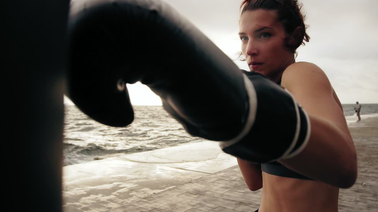 vista de cerca de una fuerte boxeadora atlética en guantes ejercitándose con una bolsa contra el hijo junto al mar. entrenamiento de boxeadora femenina