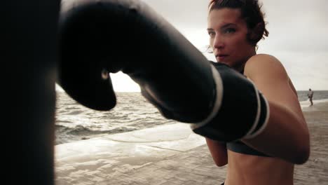 Vista-De-Cerca-De-Una-Boxeadora-Atlética-Fuerte-Con-Guantes-Haciendo-Ejercicio-Con-Una-Bolsa-Contra-El-Hijo-Junto-Al-Mar.-Entrenamiento-De-Boxeadora