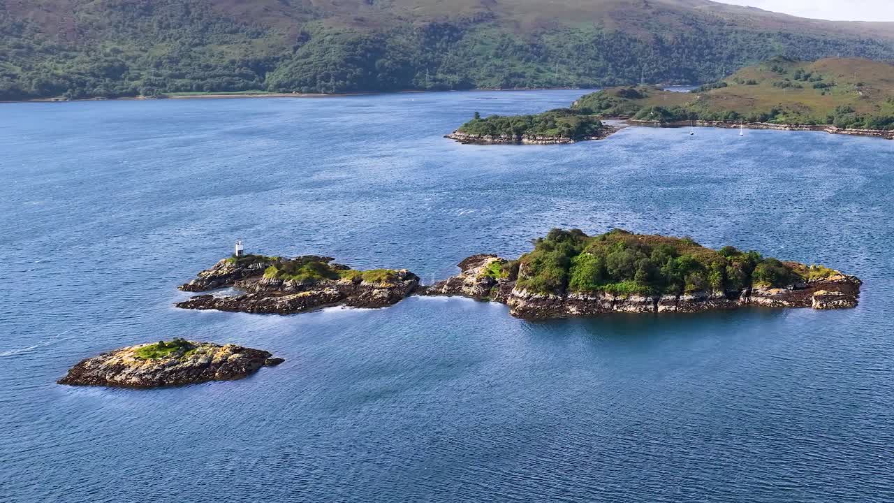 Drone glides over blue water toward green islands and lighthouse, revealing scenic Scottish coastline