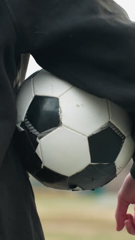 back view of someone in a black outfit holding a classic black and white soccer ball under his arm, with a blurred grassy field and buildings in the background