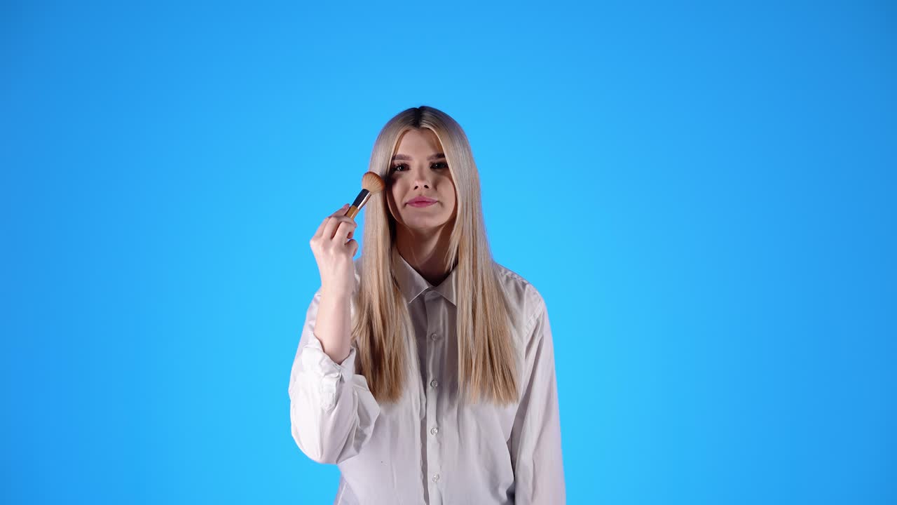 행복한 젊은 여성이 화장품 브러쉬를 잡고 메이크업을 적용합니다. 파란색 배경입니다.