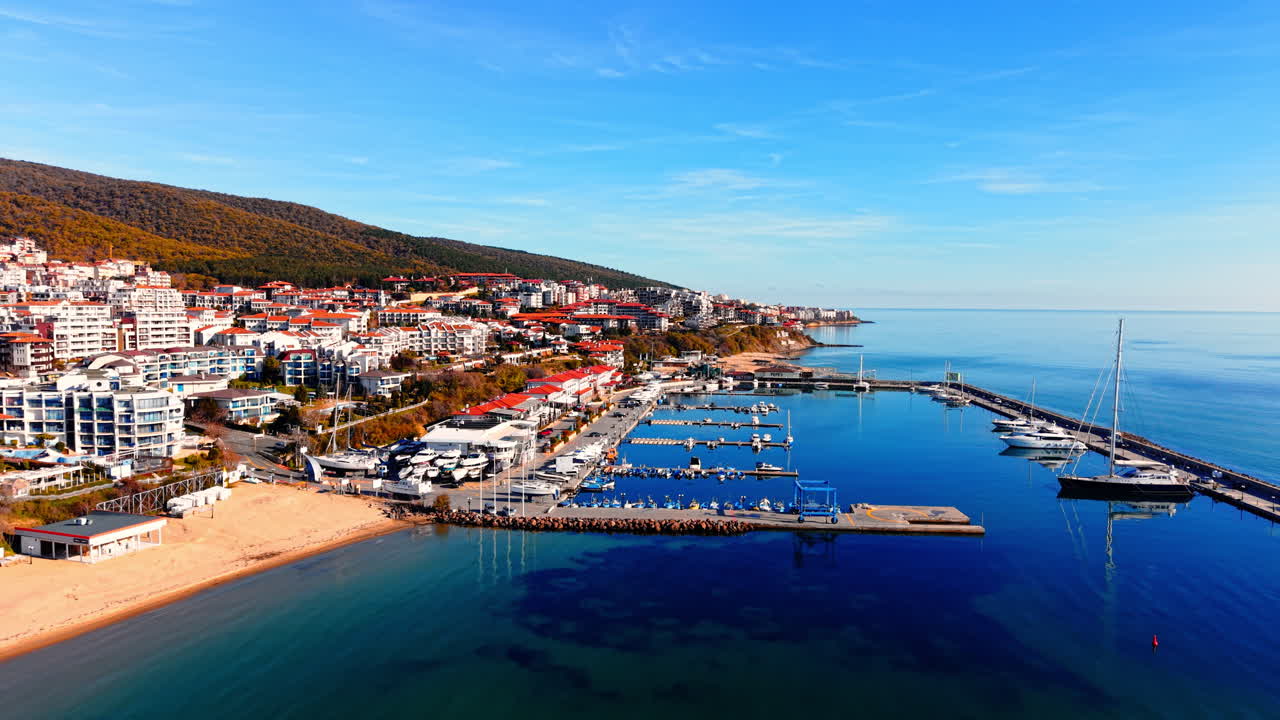 Lüks Sveti Vlas, Bulgaristan'ın havadan görünümü. Berrak mavi bir gökyüzünün altında, taş iskelede büyük yelkenli tekneler ve yatlar demirlemiş. Kırmızı çatılı binaları ve yeşil tepeleri olan kıyı kasabası
