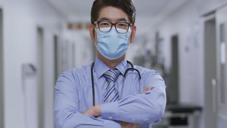 portrait of caucasian male doctor with arms crossed wearing face mask in the corridor at hospital