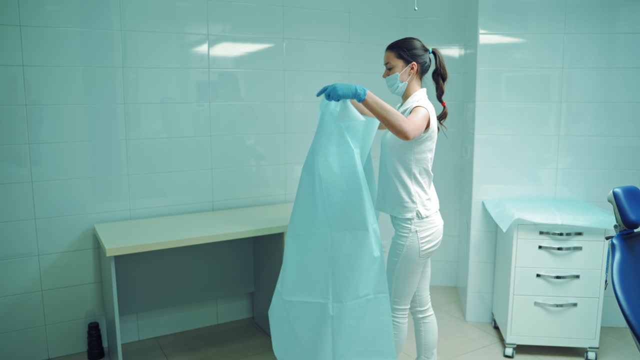 Young female nurse in white clothes lays out blue material in the hospital. Woman in mask prepares the dentist office for work day indoors.