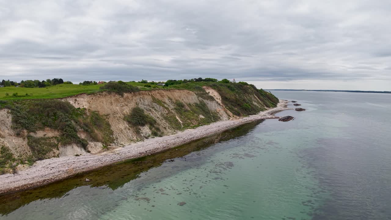 Drone aerial footage capturing rocky coastal cliffs with green vegetation along the shoreline and calm sea waters in Denmark