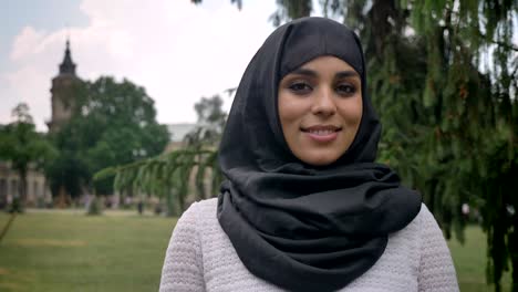 young muslim woman in hijab is smiling in daytime in park in bad weather, watching at camera, building on background, religiuos concept