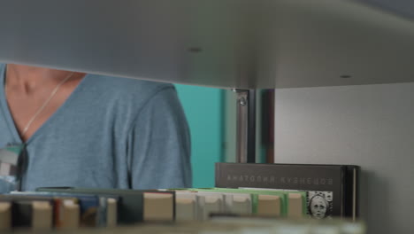 Young man in casual t-shirt and backward cap walking between bookshelves in modern library, hand resting on shelf, glancing sideways with thoughtful expression, surrounded by colorful books