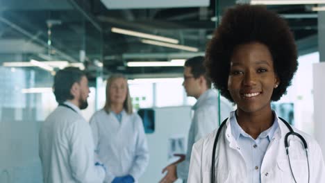 Doctora-Afroamericana-Caucásica-Mirando-Y-Sonriendo-A-La-Cámara-En-El-Consultorio-Del-Hospital