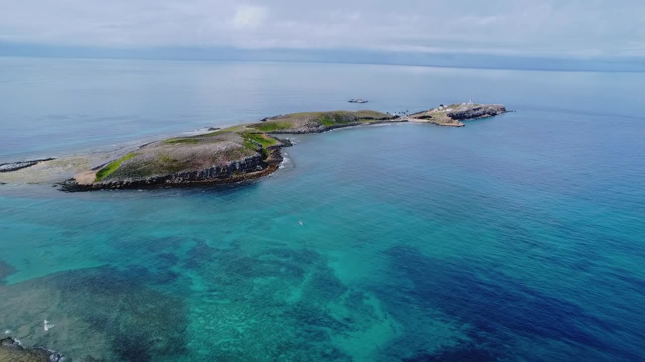 Arcipelago di Abrolhos che mostra un vasto santuario marino con acqua turchese limpida e una piccola isola con un faro distante sotto un cielo nuvoloso nell'Oceano Atlantico. Drone in avanti con zoom.