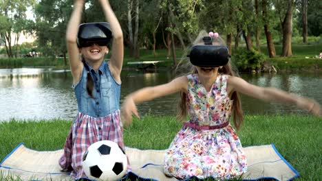 two little girls sit in virtual reality helmets on the lawn by the river and play virtual games. they clap their hands and laugh.