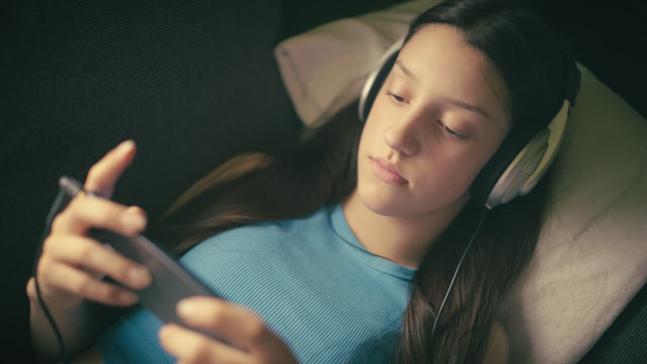Teen girl relaxing at home, listening to music with headphones and using her smartphone. A calm, intimate moment of digital leisure in a cozy setting.