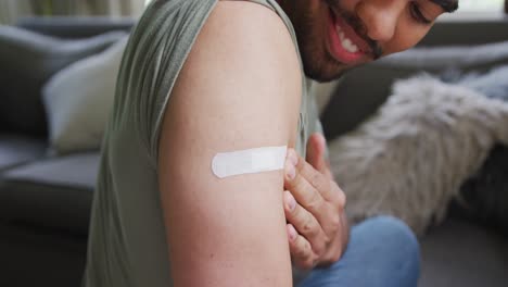 smiling young man looking at bandage on arm while sitting at home after vaccination