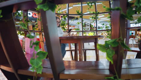 chic young woman seated in decorative wooden sit-out, holding phone, shopping bags by her side, passerby walking in vibrant, bustling mall with dynamic background lights