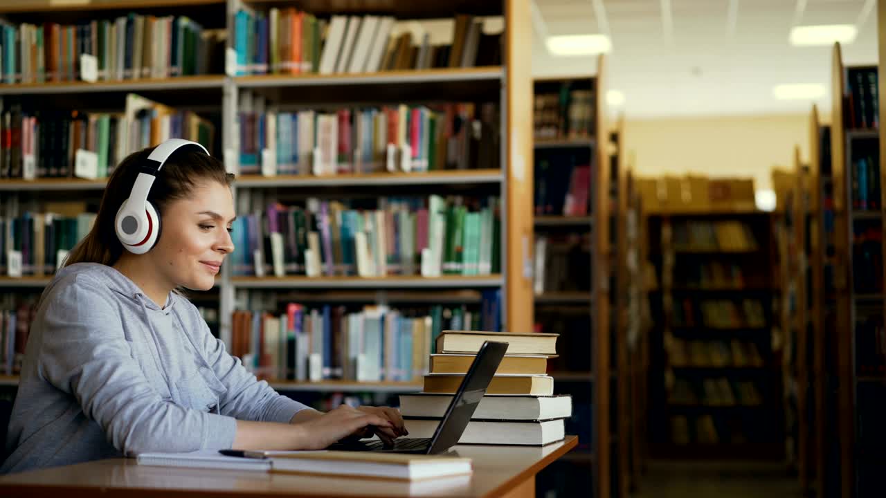hermosa estudiante caucásica positiva con grandes auriculares trabajando en la mesa en una biblioteca espaciosa frente a la computadora portátil. está sonriendo y enviando mensajes de texto