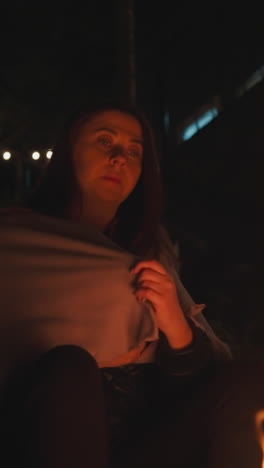 mujer aleja las manos de la llama del fuego. mujer solitaria se sienta frente a la hoguera con una manta sobre los hombros. señora calienta los dedos debido al clima frío