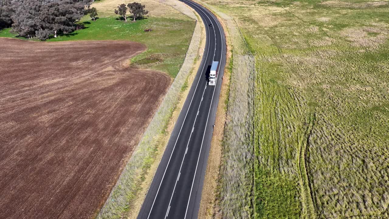 Un camion bianco viaggia lungo una strada rurale tortuosa delimitata da campi, ripreso in piena luce diurna da una prospettiva aerea alta e stabile.