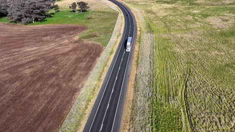 A white truck travels along a winding rural road bordered by fields, captured in bright daylight from a high, steady aerial perspective
