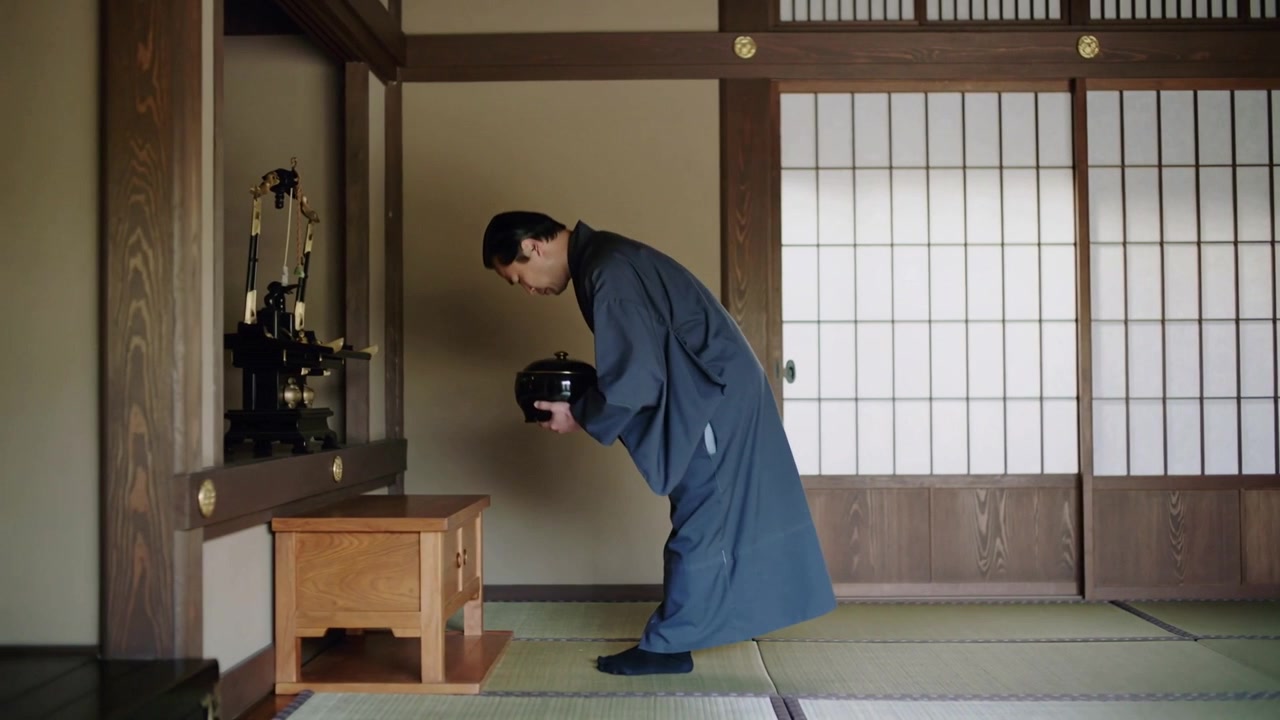 Entrando da destra, un uomo in kimono scuro cammina sul tatami, inchinandosi per presentare una ciotola laccata.
