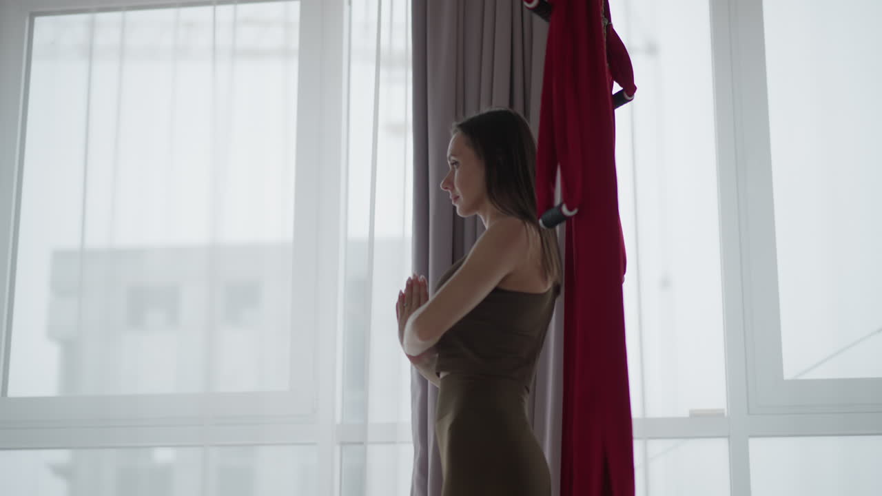 Young lady stands beside transparent window in bright hall performing yoga with hands in prayer pose next to red silk hammocks focusing on mindfulness balance serenity under soft natural light