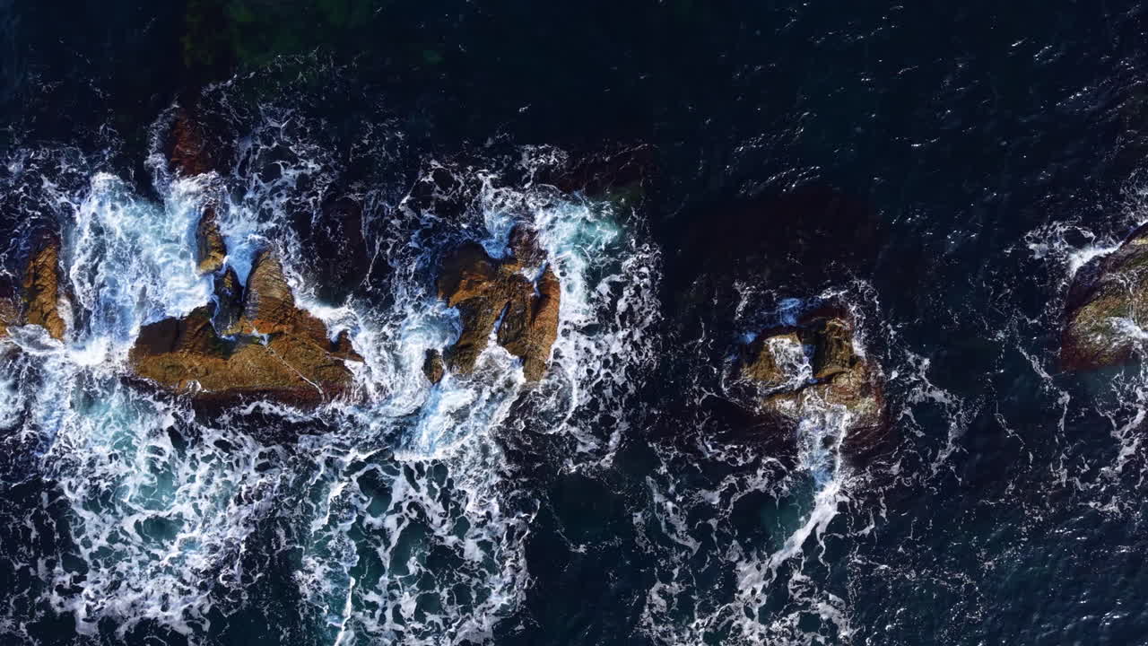 Waves crashing against rugged rocks under the vivid blue sky at midday, showcasing nature's relentless power and beauty along the coastline