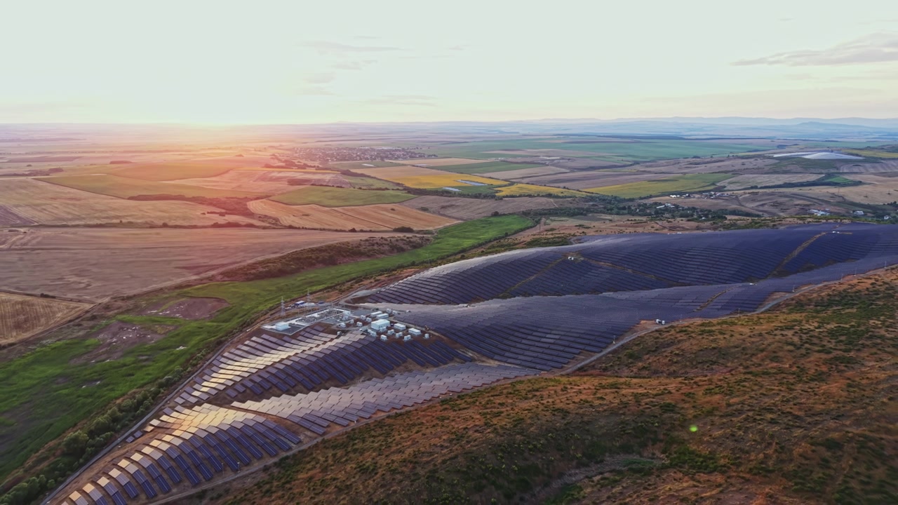 Solpaneler dækker et stort område på en bakkeskråning ved solnedgang med marker og bjerge i baggrunden.