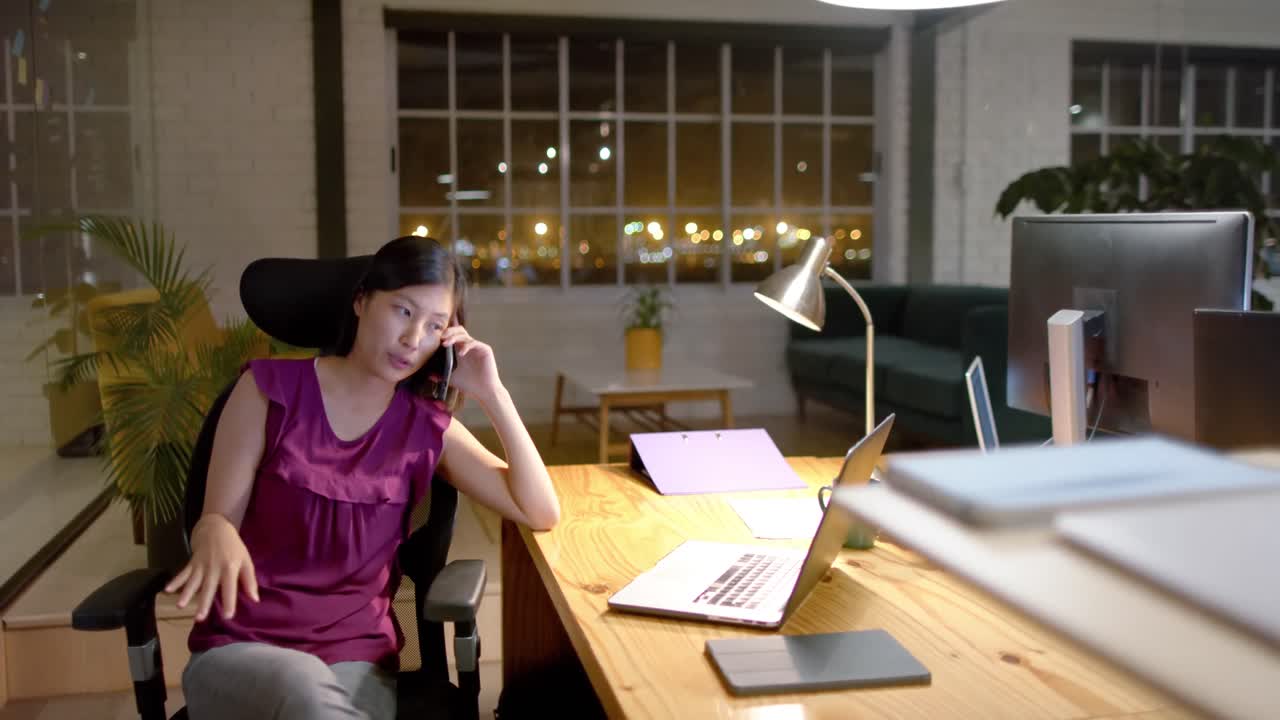 Tired asian casual businesswoman using laptop talking on smartphone in office at night, slow motion