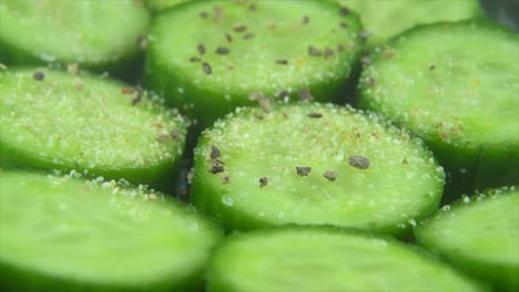 Un-Primer-Plano-Macro,-Pimienta-Que-Cae-Y-Granos-De-Especias-En-Rodajas-Cortadas-De-Un-Pepino-Verde-Salado-Fresco,-Video-De-4k-En-Cámara-Lenta