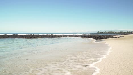 Yellow-sand-beach-with-white-capped-waves-splashing-over-lava-rock-formations-and-palm-trees-in-the-distance