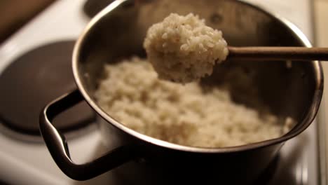 Sacudiendo-El-Arroz-Blanco-Cocido-De-Una-Cuchara-De-Madera-De-Vuelta-A-La-Olla-Metálica-En-La-Estufa,-Slomo