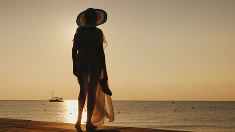 a stylish girl in fluttering clothes and a broad-brimmed hat meets the sunrise on a sea pier