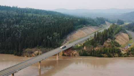 aerial drone backward moving shot over a over bridge with a car along with a camping trailer passing by the alaskan highway in usa during early morning