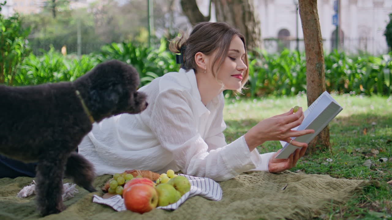 Picnic woman reading book while poodle walking blanket in park. Happy girl