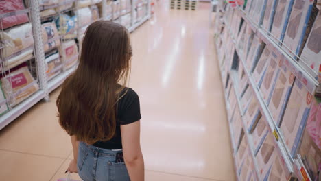 aerial perspective of elegant young woman in black top and glasses walking through store aisle, browsing goods on shelves while looking around, bright retail lighting and organized supermarket