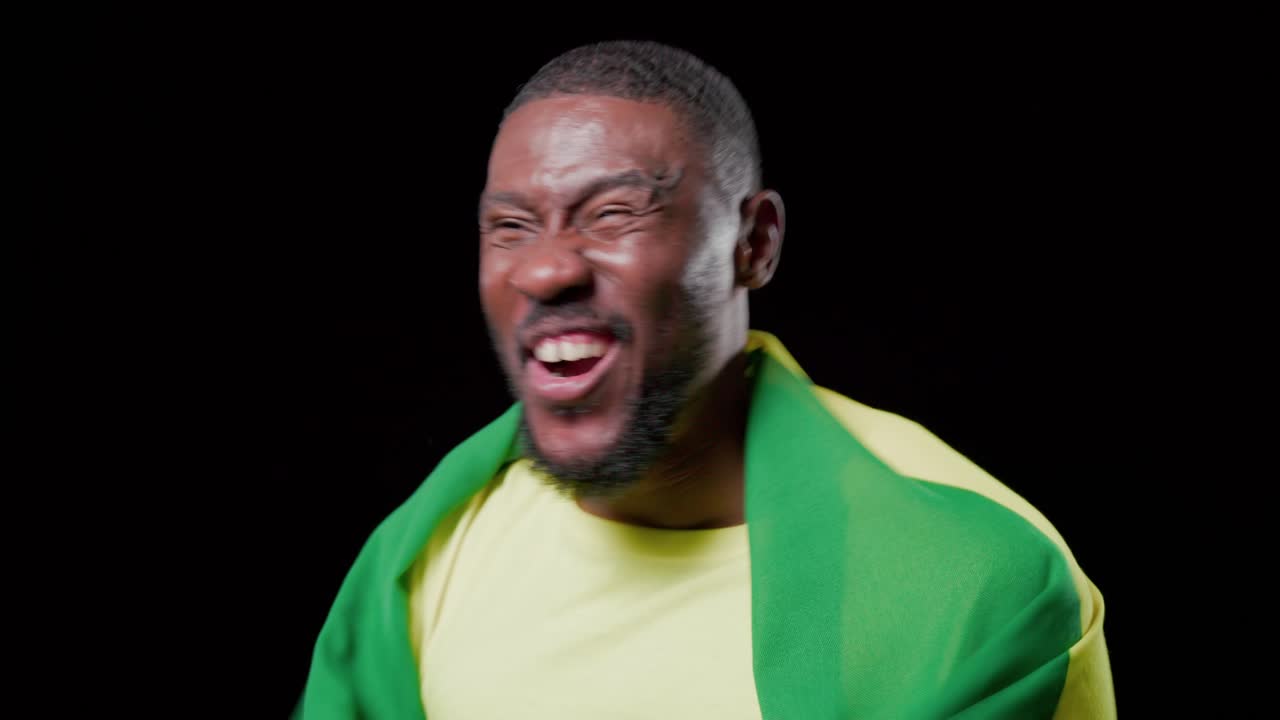 Mid adult man shouting, lifting and waving Brazil flag on black backdrop, yellow tee, celebrating
