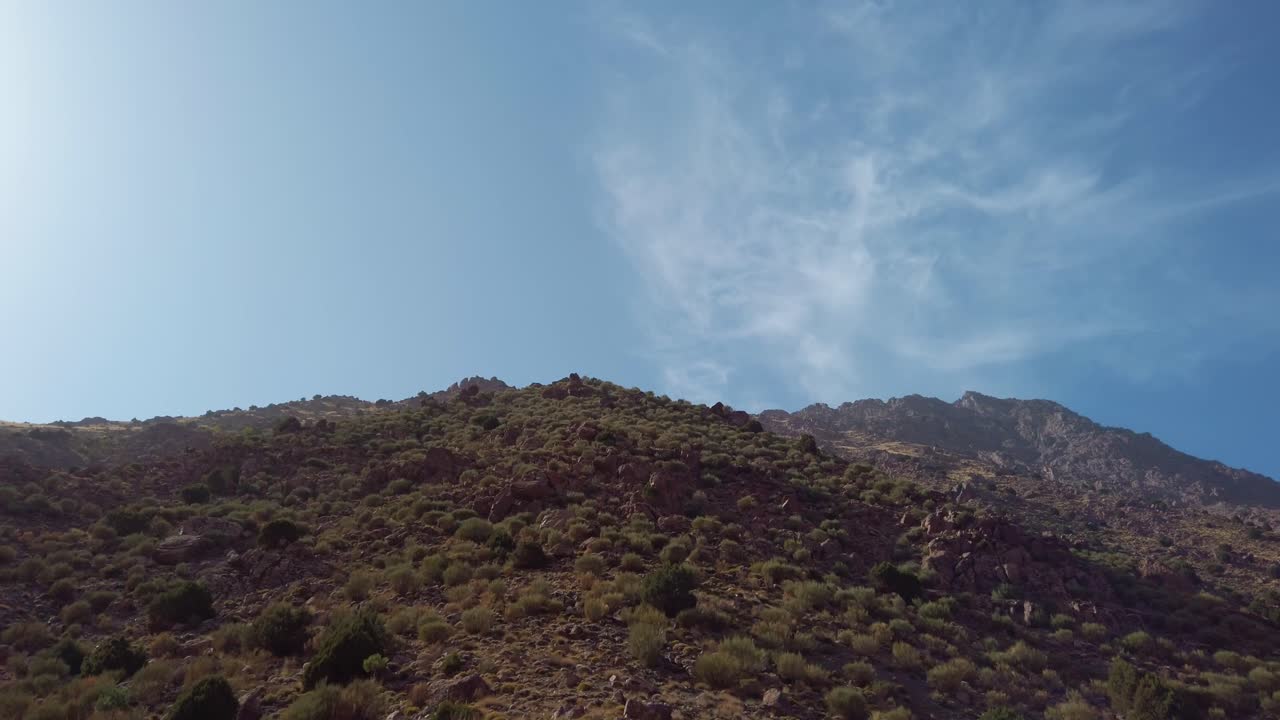 Vista dei pendii rocciosi delle montagne con arbusti sparsi nelle montagne dell'Alto Atlante, vicino a Imlil, Marocco, sotto un ampio cielo blu con nuvole sottili, che evidenzia il paesaggio naturale aspro del Monte Toubkal