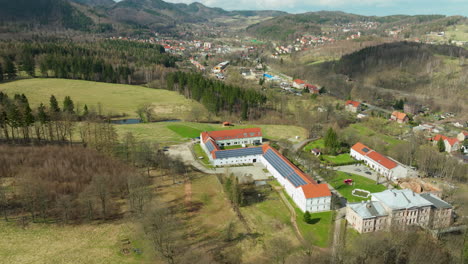 An-aerial-view-of-a-large-building-with-an-orange-tiled-roof-surrounded-by-a-serene-landscape-with-lush-trees,-a-tranquil-pond,-and-a-backdrop-of-a-quaint-town-nestled-in-the-rolling-hills