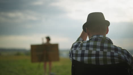 a back view of a man wearing a black hat, adjusting it as he sits in a grassy field. in the background, a blurred figure of a woman is painting, creating a outdoor artistic scene under a cloudy sky