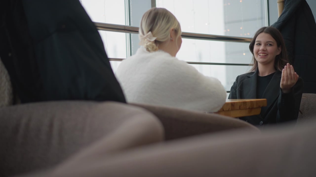 Due donne sono sedute una di fronte all'altra a un tavolino accanto alla finestra all'interno di un luminoso interno moderno, condividendo una conversazione calda e attenta con gesti rilassati e luce diurna soffusa.