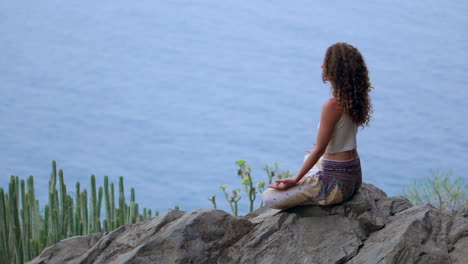 positioned on a mountaintop rock on an island, a young woman engages in yoga, meditating in lotus position, gazing at the ocean