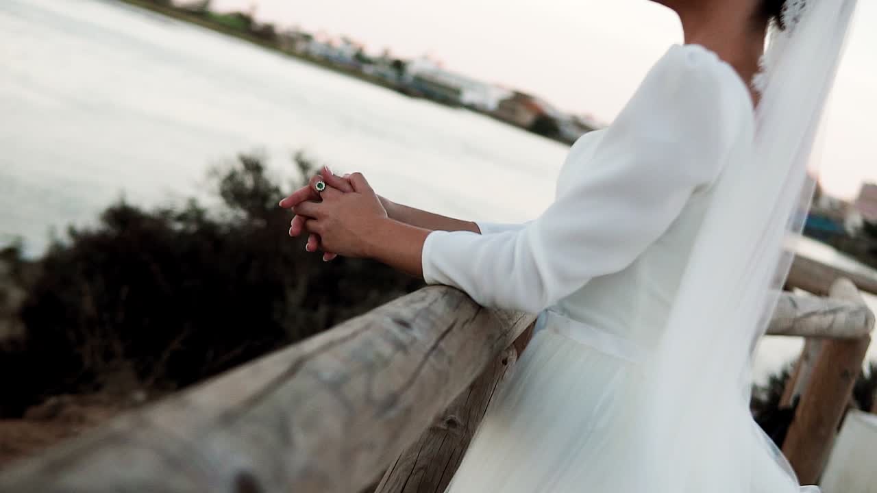 Sposa elegante in abito bianco appoggiata a una ringhiera di legno di fronte a un lago al tramonto