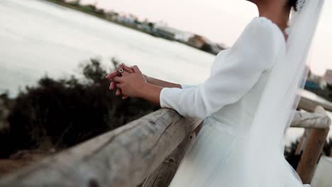 elegant bride with white dress leaning on a wooden railing in front of a lake at sunset