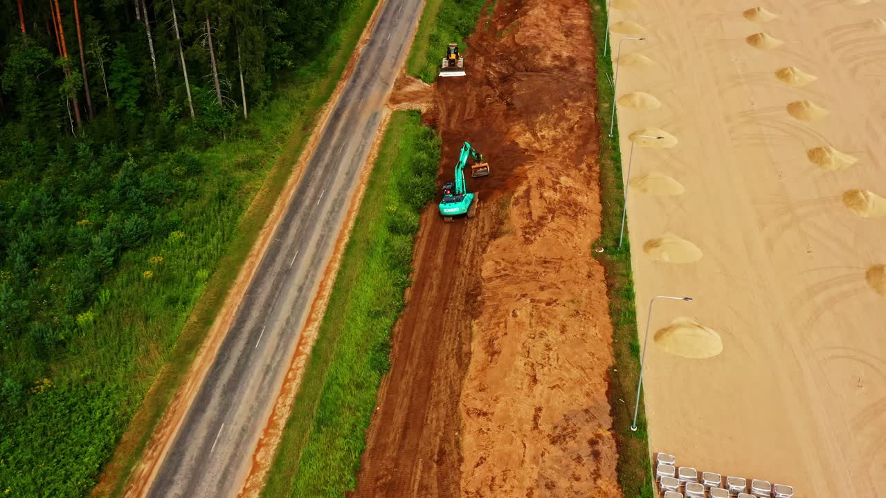 Excavator Working At Construction Site Between Rural Road And Sandy Terrain With Stacked Concrete Blocks. aerial flyby shot