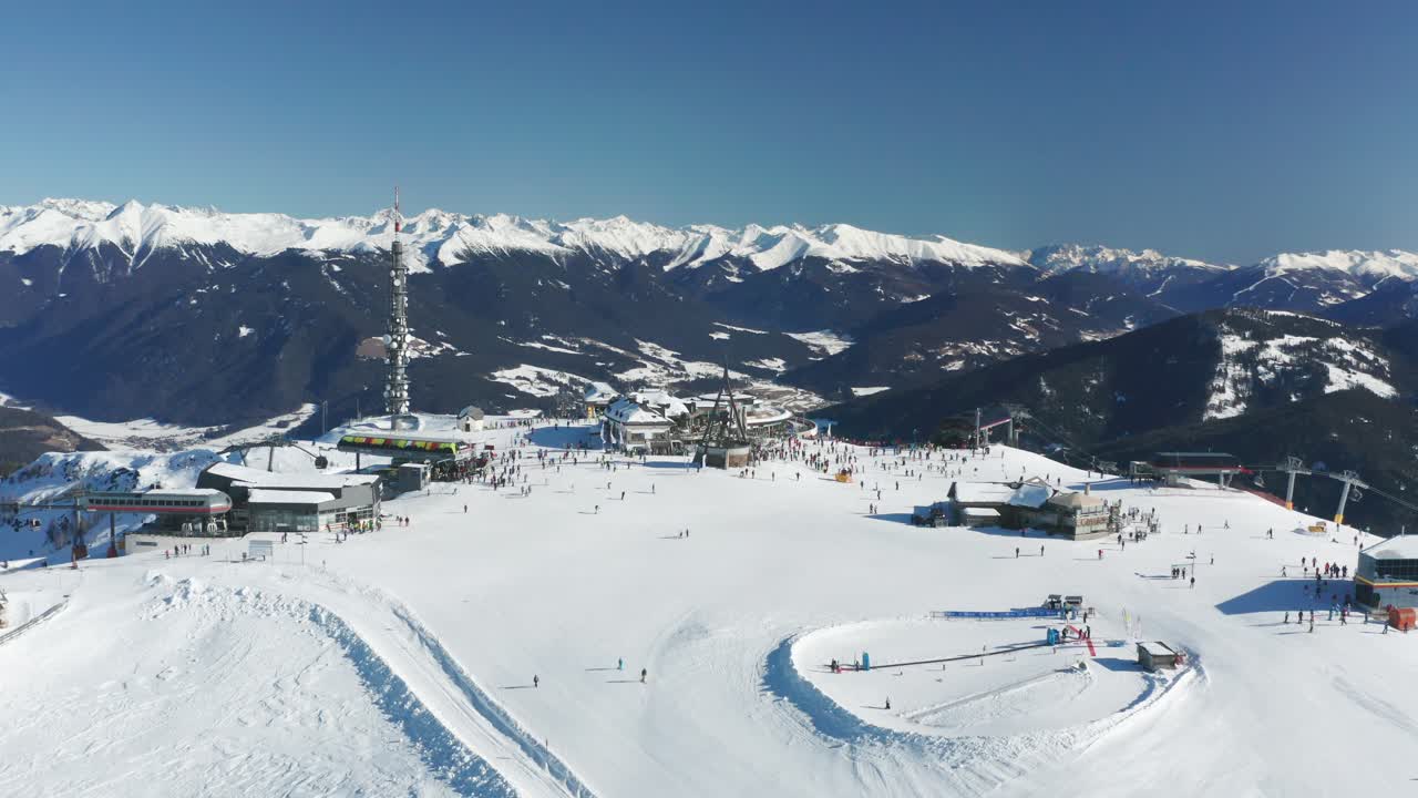 Aerial view of the Kroplatz mountain top, a famous skiing resort in Italy. Concordia bell is ringing, skiers ride on the plateau. It is sunny, the sky is clear, the valley below is covered with snow.