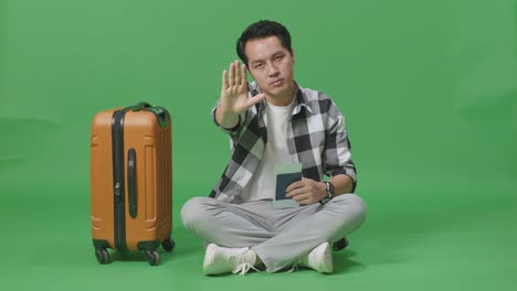 full body of asian male traveler with luggage and passport looking at camera and disapproving with no hand sign in the green screen background studio