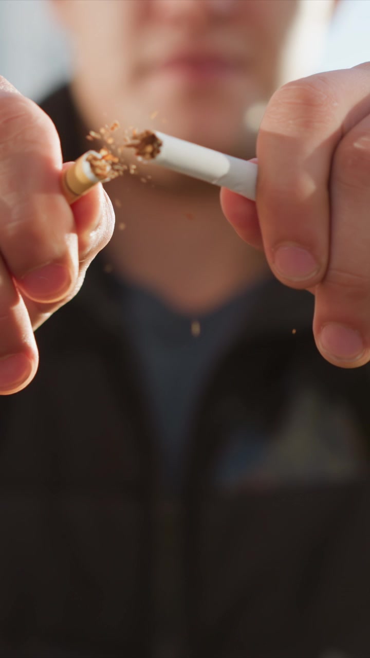 Primer plano de un hombre sosteniendo y encendiendo un cigarrillo, con detalles visibles de la mano y la punta encendida. El fondo muestra un entorno urbano con edificios desenfocados que crean profundidad. La iluminación resalta la piel.