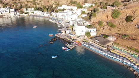 drone view in greece flying over blue sea in loutro small white house town and small boats next to a hill on a sunny day