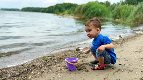 caucasian toddler plays with bucket and shovel on the shore. happy child spending time at the water on windy summer day.