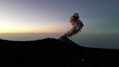 drone view in guatemala flying over a volcano crater with an erupting volcano at sunrise and shadow of people on the rim