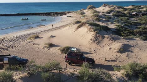 Toma-De-órbita-Aérea-Vehículo-4x4-Conduciendo-Desde-Una-Playa-De-Arena-Con-Océano-En-Dirección-Camino-Rural-Durante-La-Puesta-De-Sol