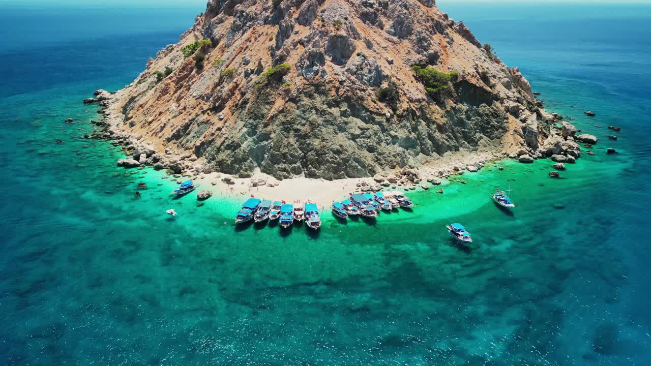 Révélation aérienne par drone de l'île de Suluada en Turquie, montrant une colline rocheuse s'élevant au-dessus d'une plage de sable avec des eaux méditerranéennes turquoise et des bateaux de touristes ancrés le long du rivage